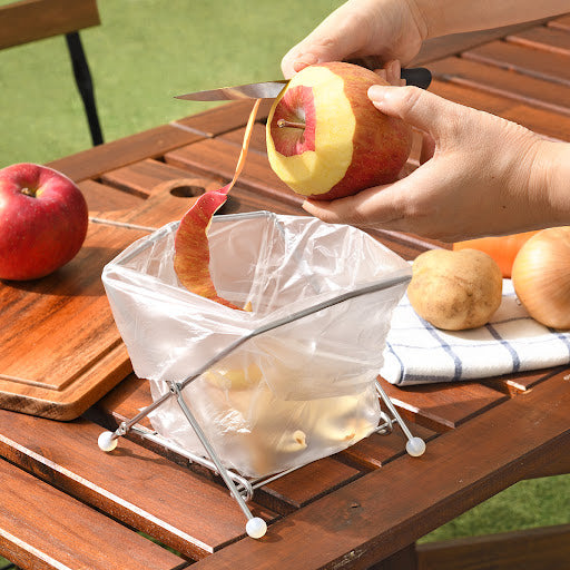 A person slices an apple into pieces on a table, with an EcoFrame Stainless Compost Holder nearby for easy disposal.