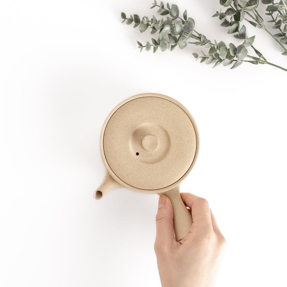 Top-down view of the beige teapot with a hand on the handle, highlighting its compact size and ergonomic design.