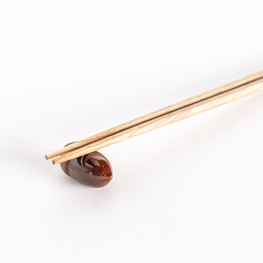 A close-up of a ceramic acorn chopstick rest holding a pair of chopsticks, showcasing its glossy brown finish and textured top.