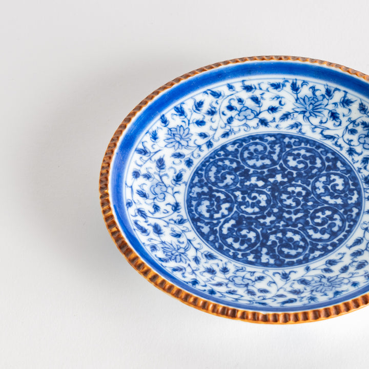 A ceramic plate with an intricate blue floral design centered around a bold circular pattern, framed by a copper-colored textured rim.