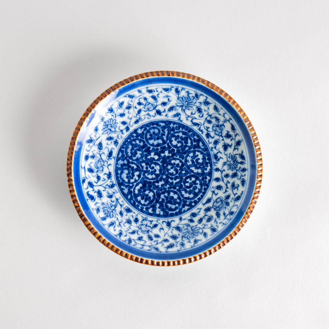 A ceramic plate with an intricate blue floral design centered around a bold circular pattern, framed by a copper-colored textured rim.