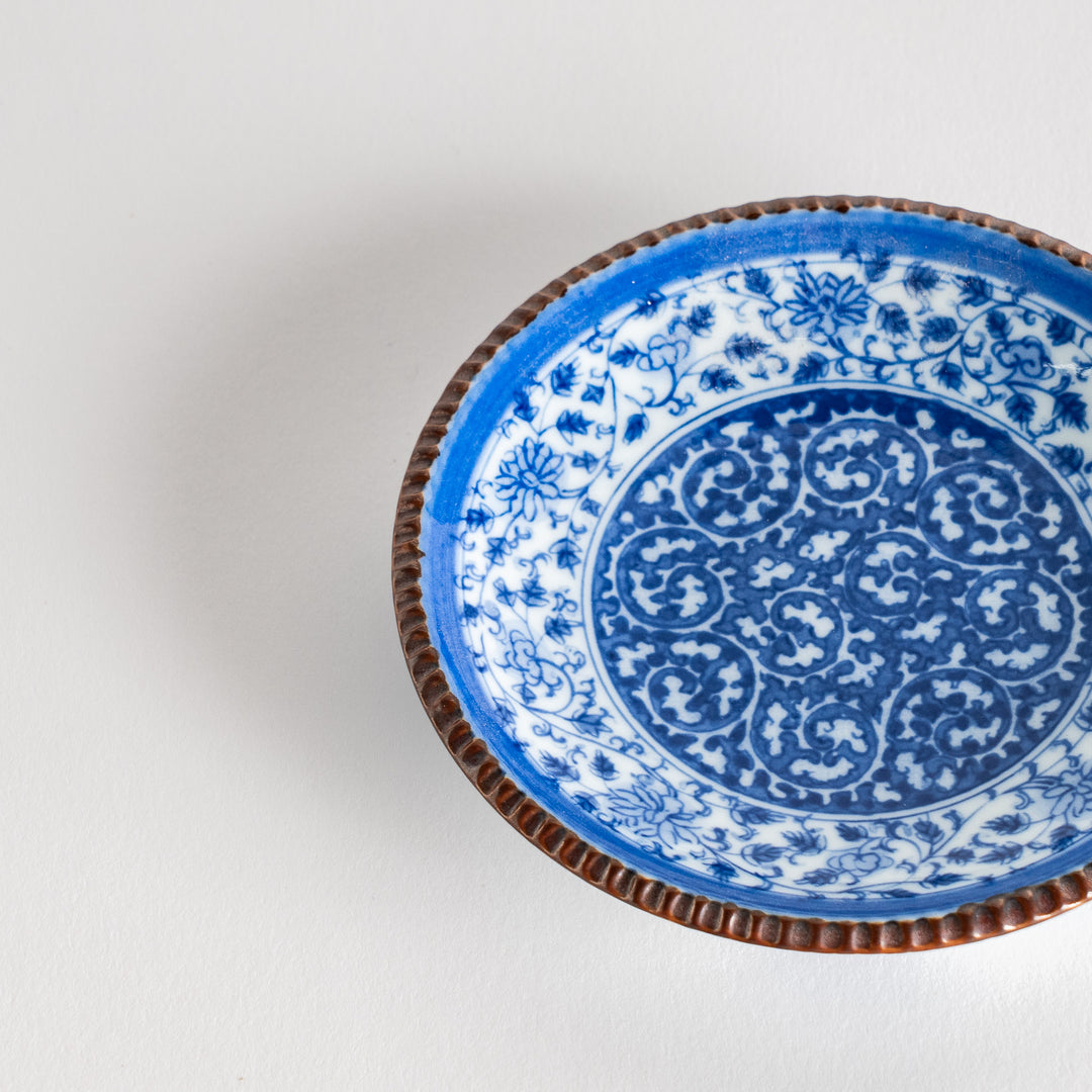 A small ceramic plate with an intricate blue floral design centered around a bold circular pattern, bordered by a copper-colored textured rim.