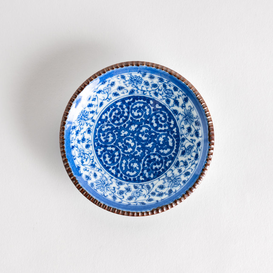 A small ceramic plate with an intricate blue floral design centered around a bold circular pattern, bordered by a copper-colored textured rim.