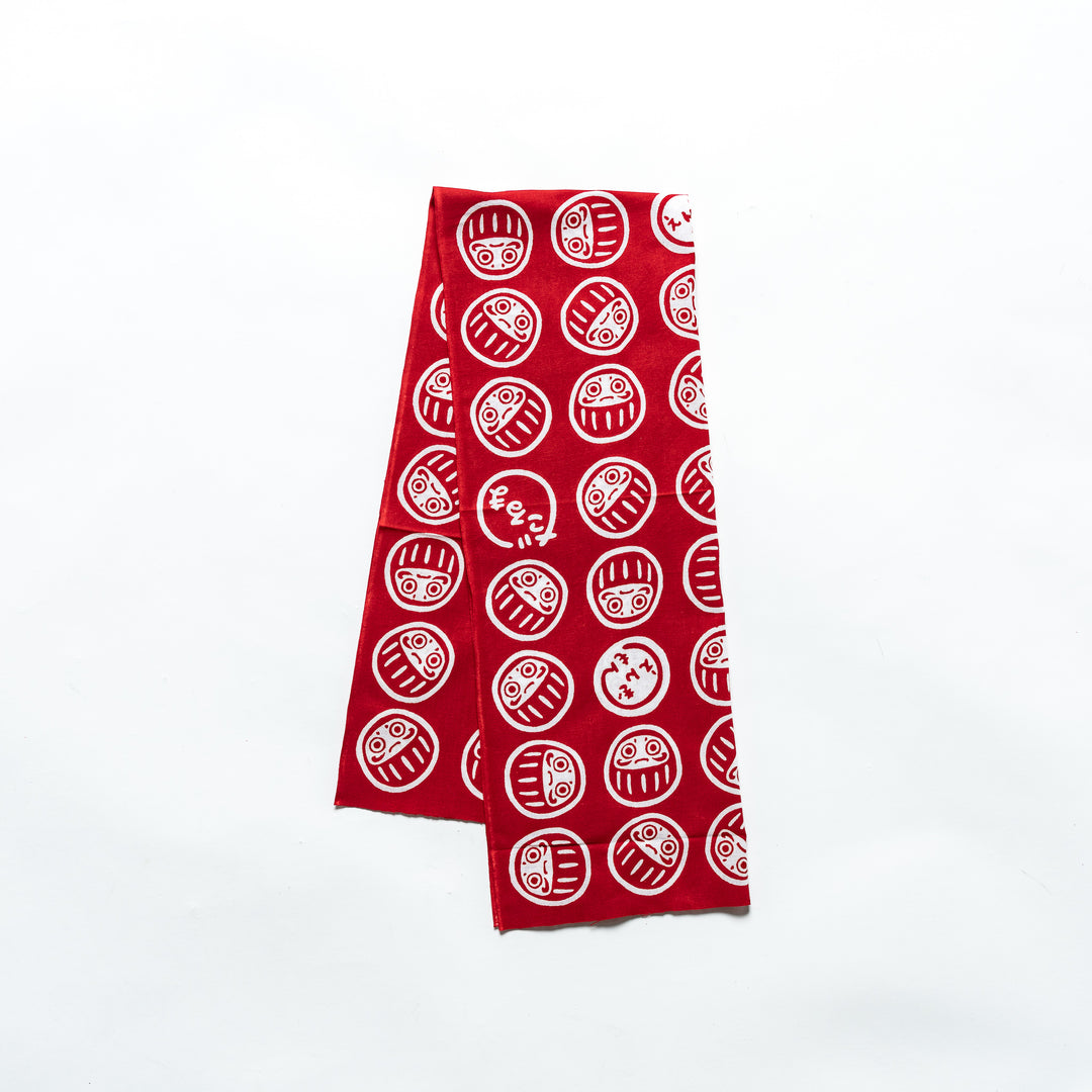 Red tenugui featuring white Daruma patterns, neatly folded, displayed on a plain white background.