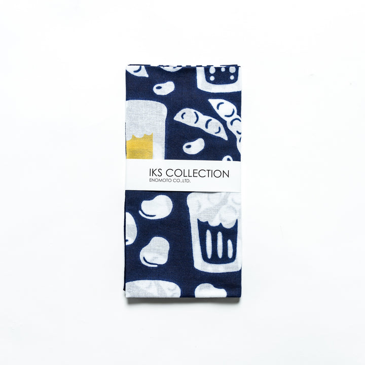 Dark navy tenugui folded with visible product sleeve featuring the beer mug and edamame motif.