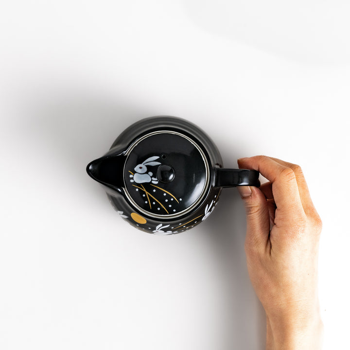 Hand holding the handle of a black teapot featuring white rabbits and golden grass, with a lid showcasing a playful rabbit design.