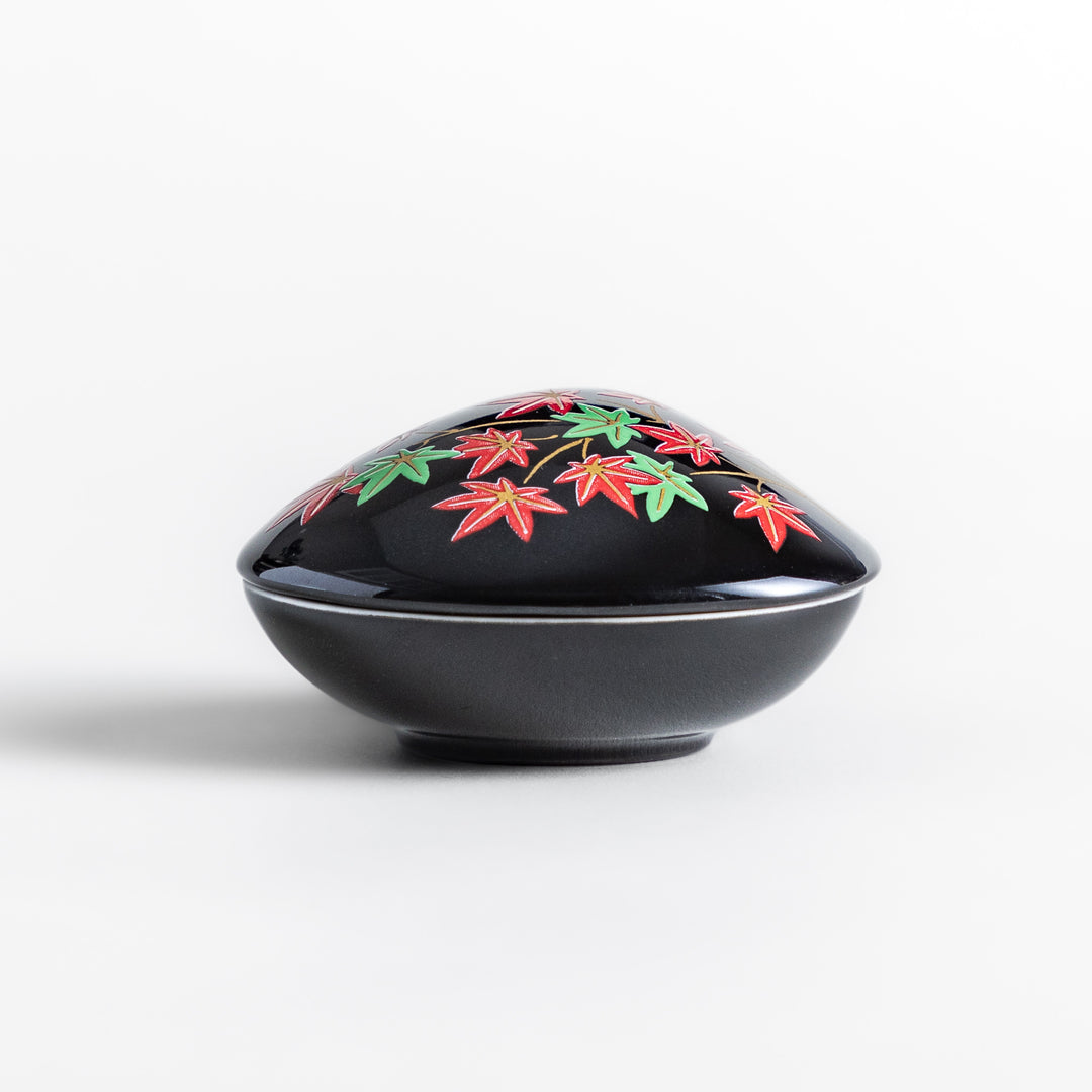 Black ceramic incense holder shaped like a seashell, adorned with red and green maple leaves, featuring a glossy red incense stand inside for secure placement.