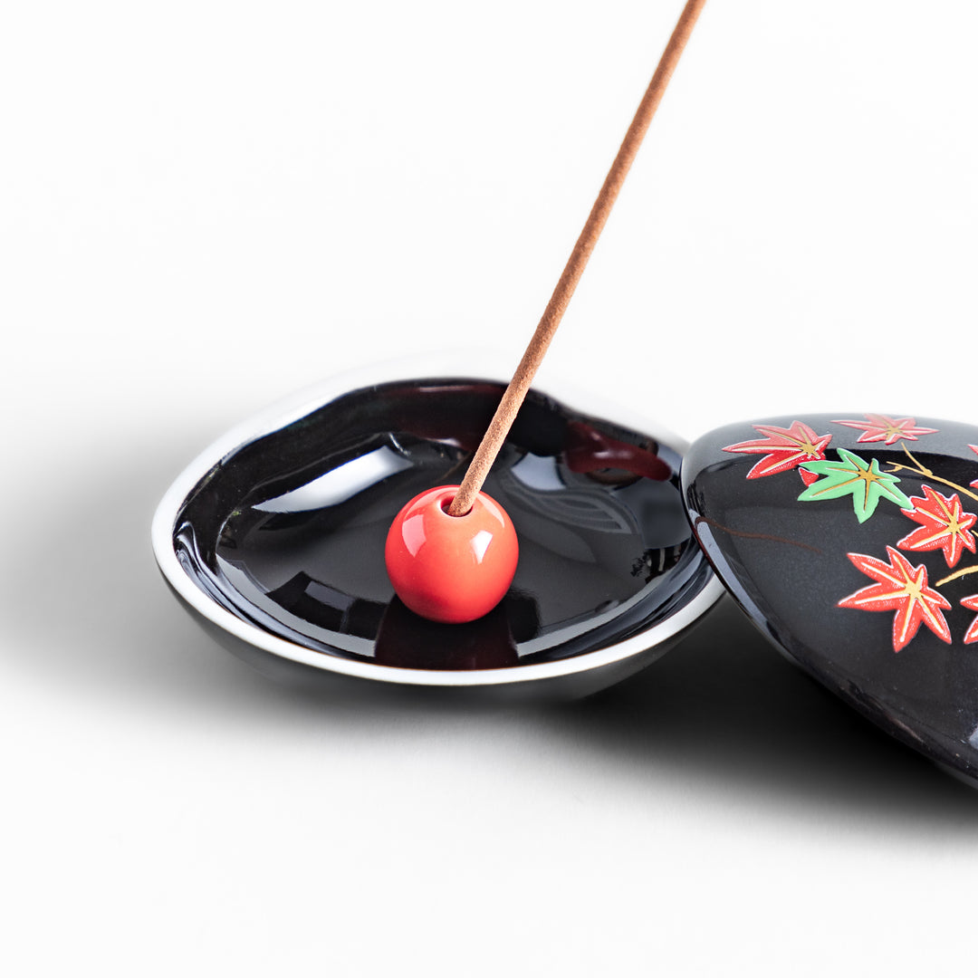 Black ceramic incense holder shaped like a seashell, adorned with red and green maple leaves, featuring a glossy red incense stand inside for secure placement.