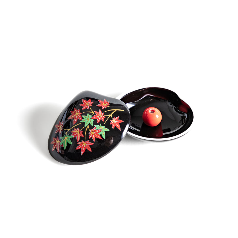 Black ceramic incense holder shaped like a seashell, adorned with red and green maple leaves, featuring a glossy red incense stand inside for secure placement.