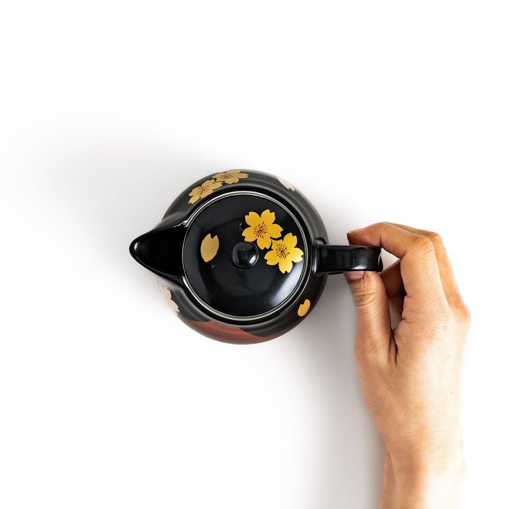 Top view of a black teapot held by hand, featuring yellow sakura blossoms and a vibrant red Mount Fuji design for added style.