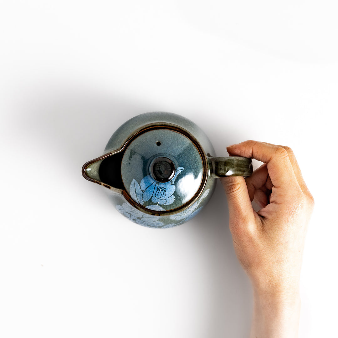 Hand gently holding the handle of a blue ceramic teapot with a lid, adorned with a delicate white camellia flower pattern.