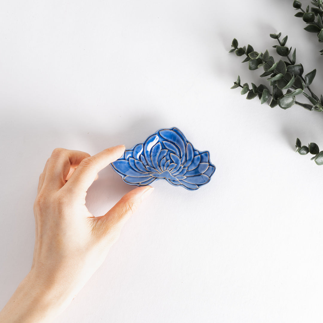 Hand holding the blue mini sauce dish, which features a gold chrysanthemum design, with decorative greenery in the background.