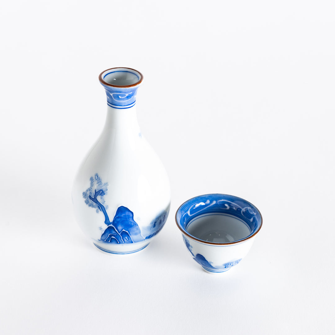 A blue and white sake set featuring a sake bottle and matching cup with hand-painted mountain and tree motifs.