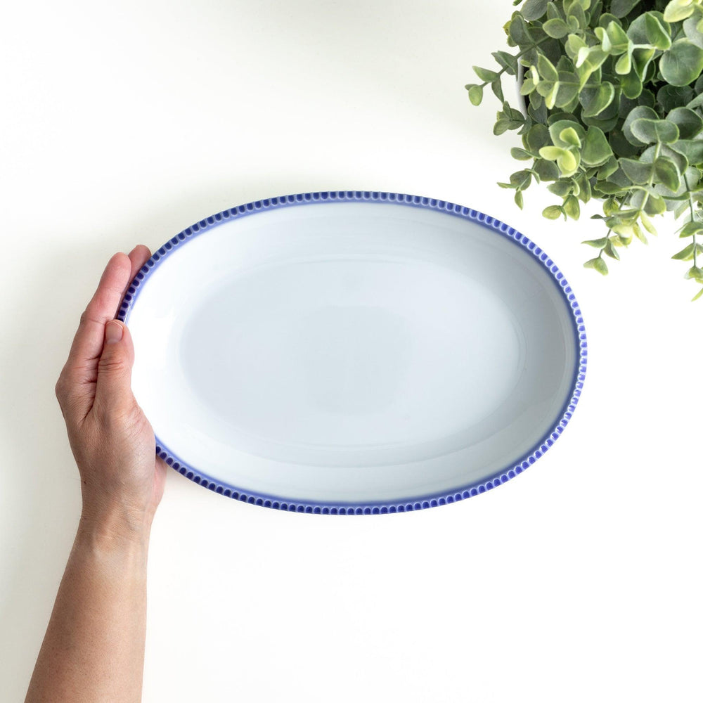 An oval-shaped white dinner plate with a rustic scallop-edged design. The border comes in blue, brown, and olive.