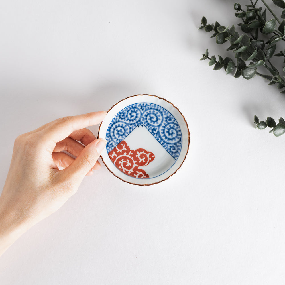 A hand holds this raised rim sauce dish featuring blue spirals and red clouds beneath Mount Fuji, blending tradition with elegance.