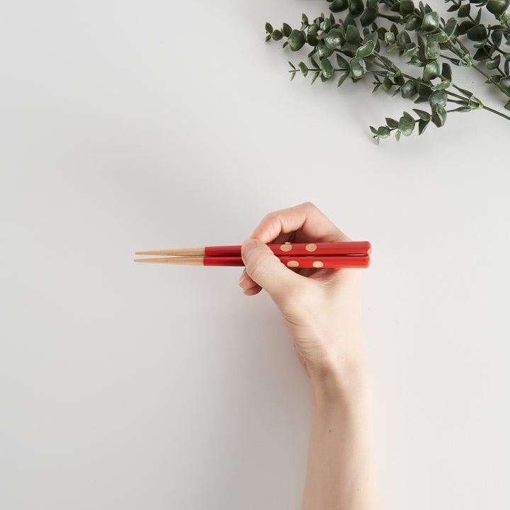 A hand holding red children's chopsticks with beige dots, positioned above a plain background.