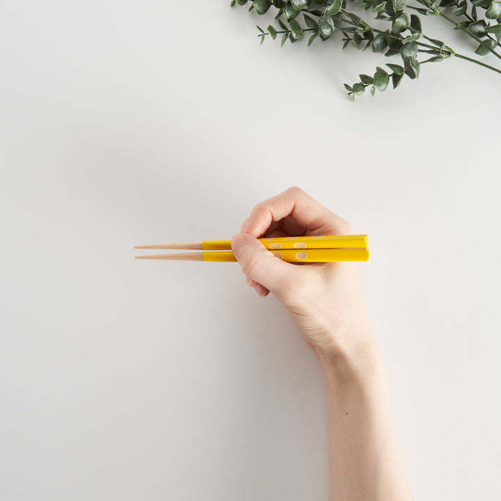 A hand holding yellow children's chopsticks, demonstrating the proper grip, with some greenery in the background.