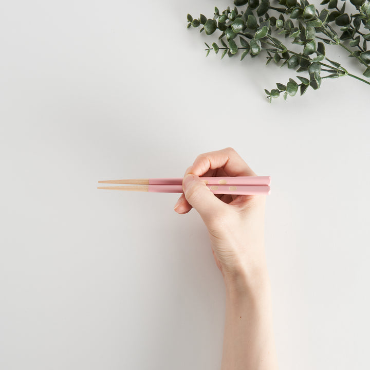 Hand holding pink children’s chopsticks with dot accents and wood tips, with green plant in the background.