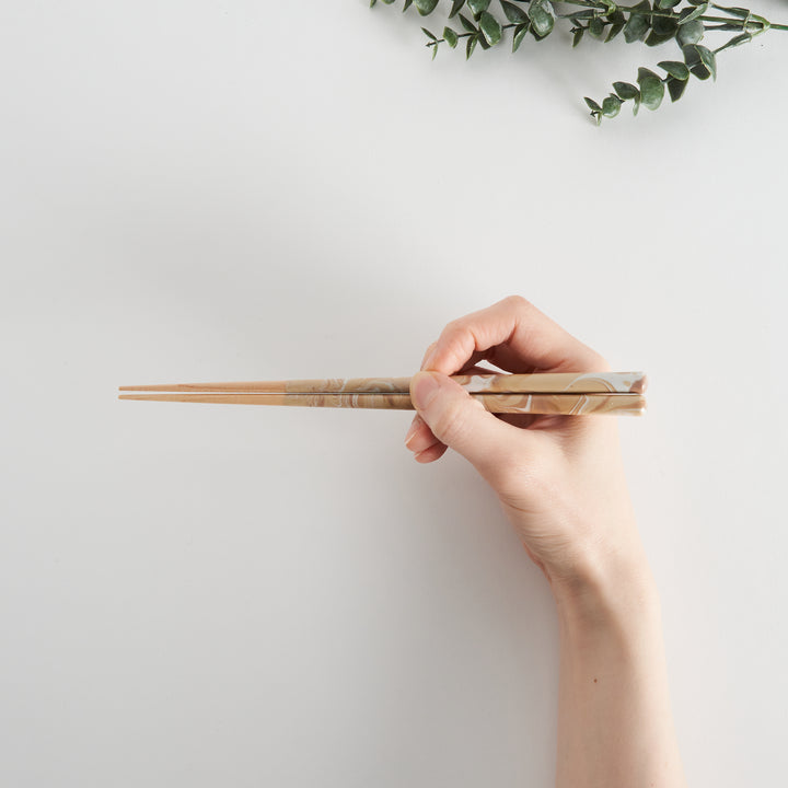 Hand holding caramel-patterned chopsticks against a light background, with eucalyptus leaves in the corner.