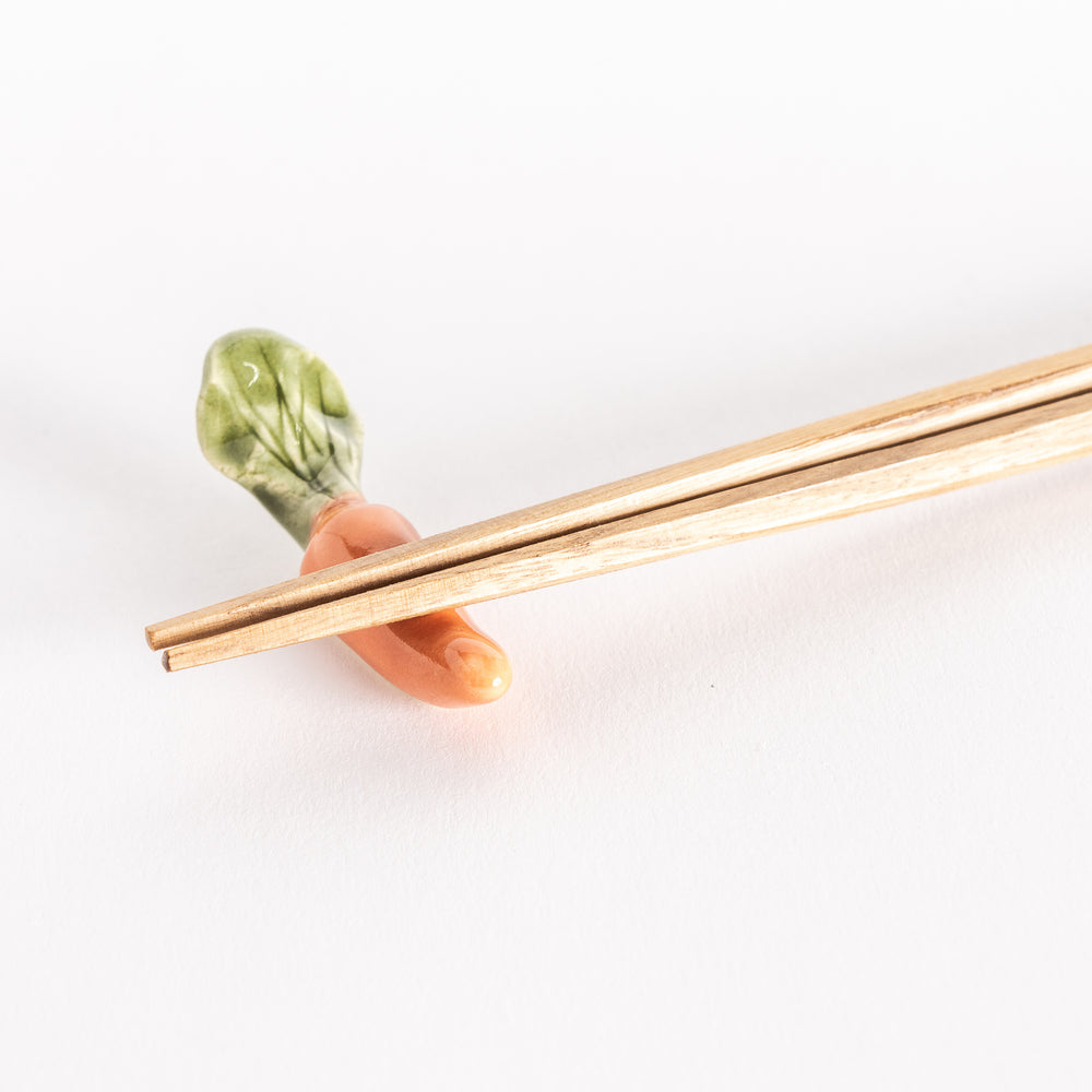 Close-up of carrot-shaped ceramic chopsticks rest with chopsticks placed, showing realistic green leafy top.