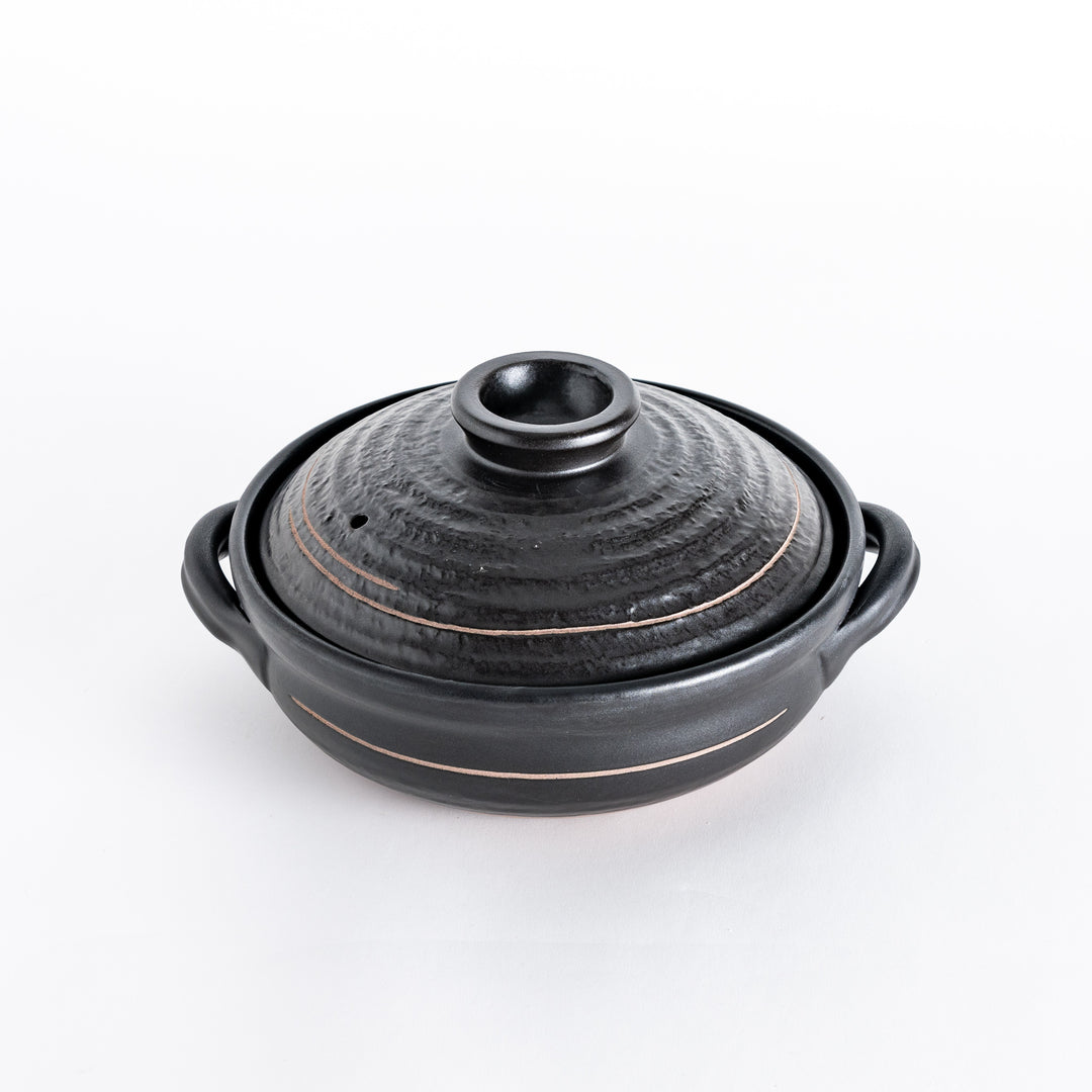 Black ceramic donabe with textured lid and two side handles, placed on a white surface.