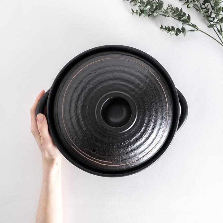 Hand holding the donabe, viewed from above, showing the top of the textured black lid.