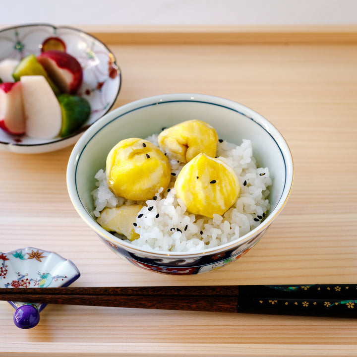 Top view of a bowl of chestnut rice, garnished with black sesame seeds, served with a side of pickled vegetables and chopsticks.