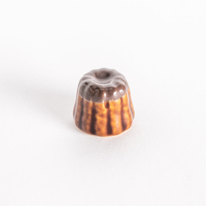 A ceramic chopstick rest shaped like a chocolate canelé, featuring a glossy dark top and ridged sides on a white background.