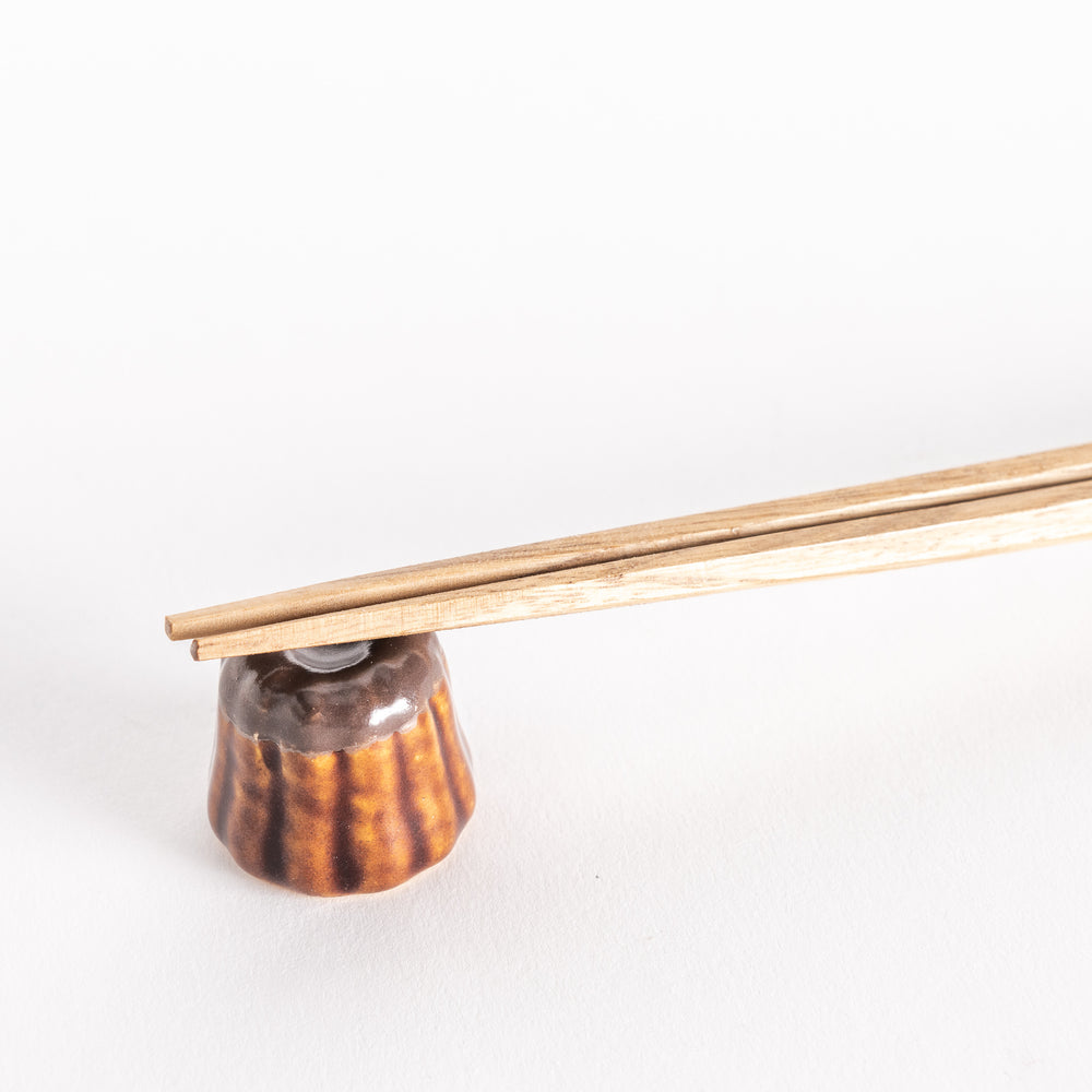 A close-up of a chocolate canelé-shaped ceramic chopstick rest holding chopsticks, showcasing its detailed texture and glossy finish.