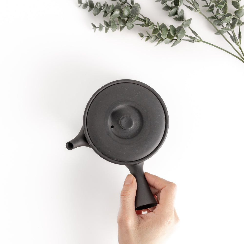 Top-down view of the black teapot with a hand on the handle, showcasing its ergonomic design and practical proportions.