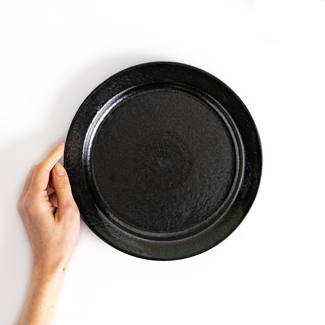 Black ceramic dinner plate held in hand, showcasing its size. The glossy surface highlights its simple and sophisticated design.