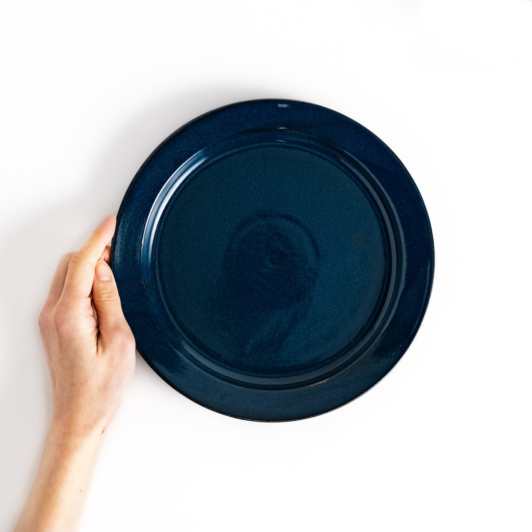 Hand holding a blue ceramic dinner plate with a glossy finish, reflecting its deep and elegant tone.