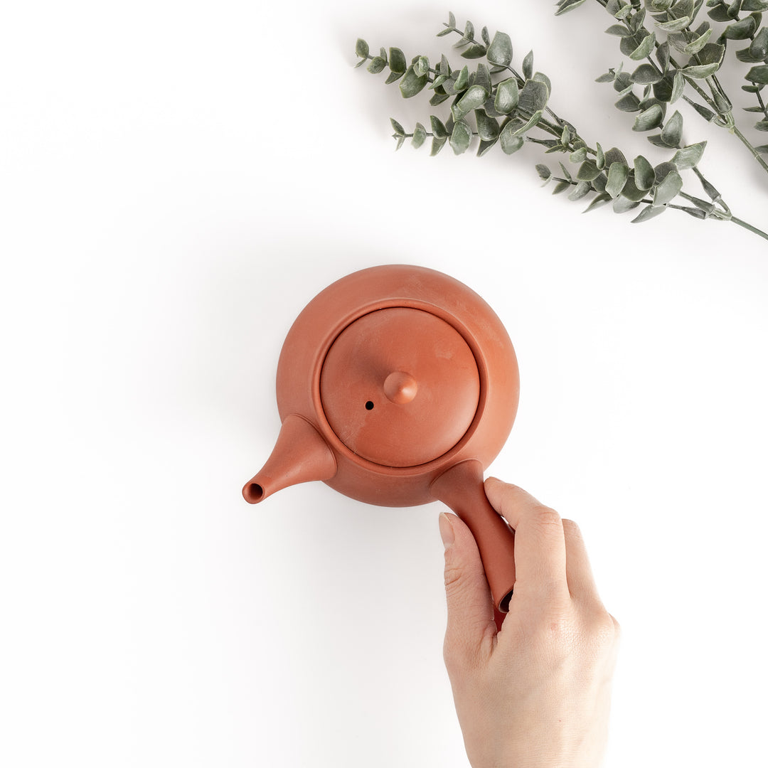 Top-down view of the crimson teapot, with a hand gently holding its handle, showcasing its balanced and ergonomic design.