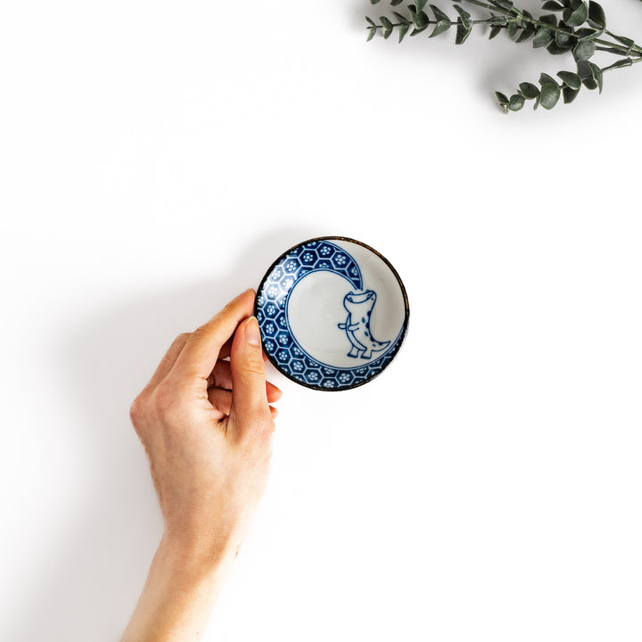 Hand holding a small sauce plate with a cute dinosaur design and a traditional Japanese geometric pattern, perfect for serving soy sauce or other condiments.