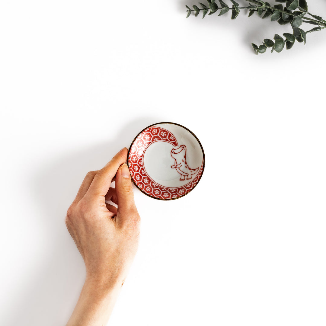 Hand holding a small sauce plate with a cute dinosaur design and a traditional Japanese geometric pattern, perfect for serving soy sauce or other condiments.