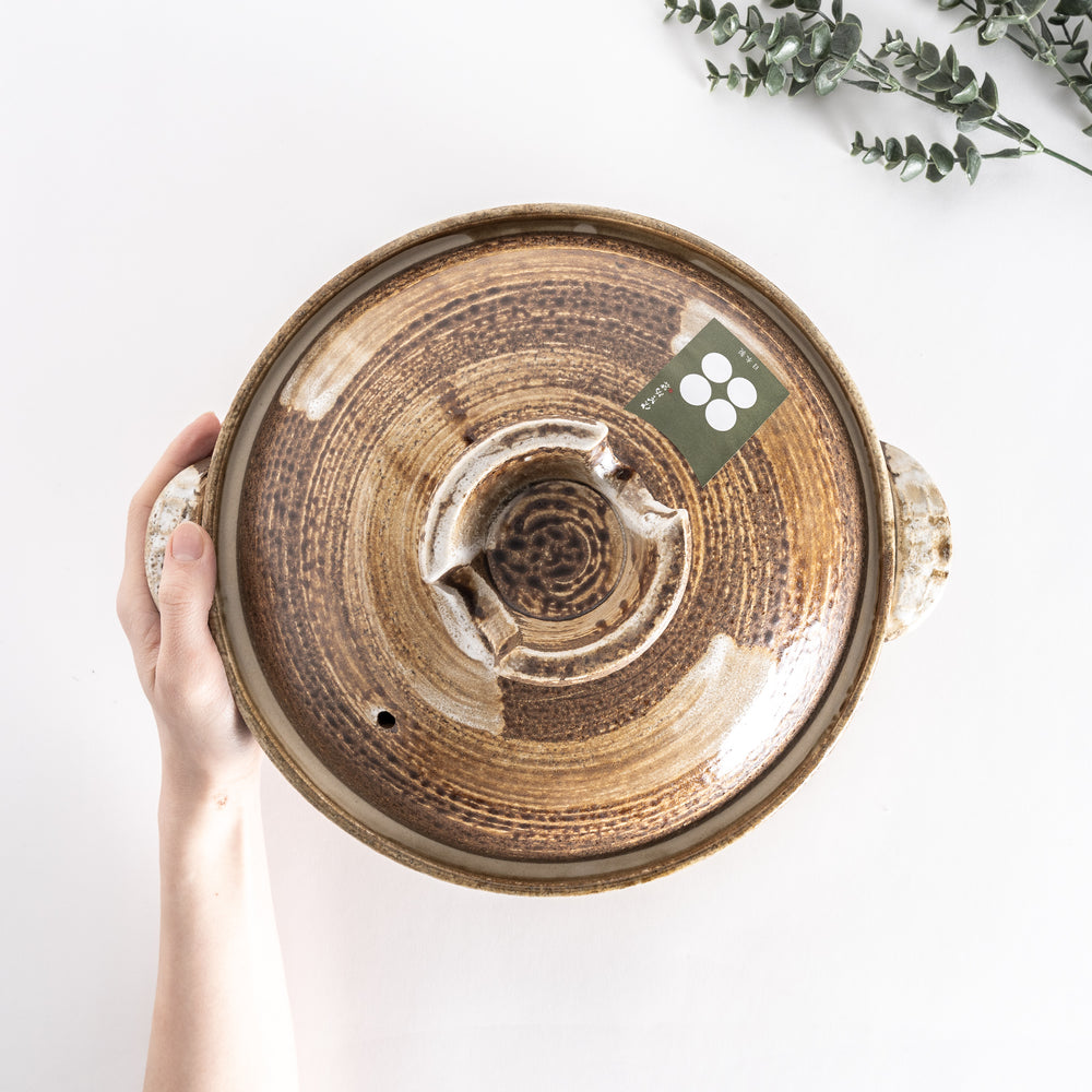 Top-down view of the donabe held by hand, showcasing the brown swirling glaze and circular handle on the lid.