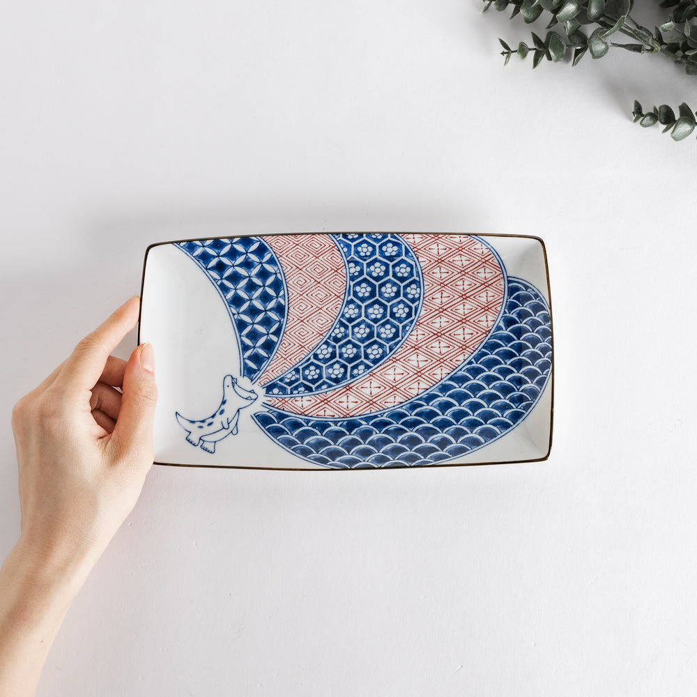 Hand holding the rectangular dessert plate with a dinosaur design, showing intricate blue and red patterns.