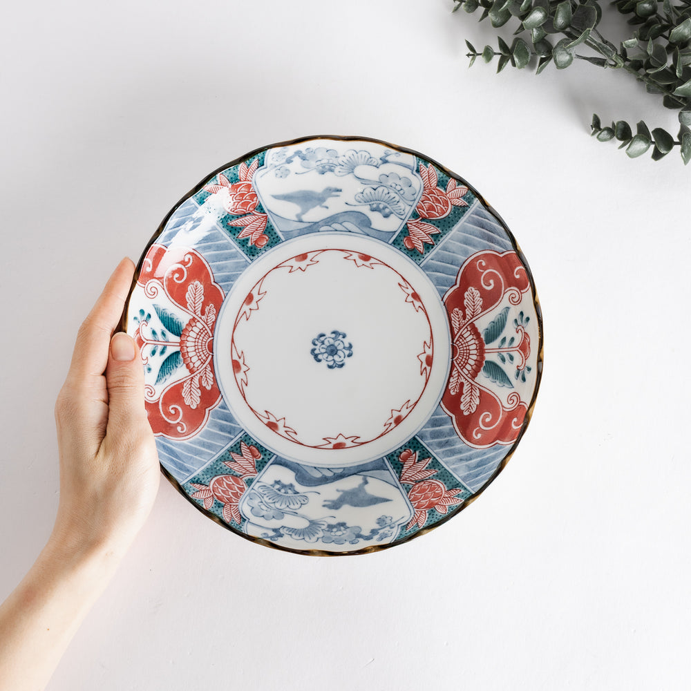 A hand holding the decorative plate, showcasing the detailed artwork in red, blue, and green against a white background.