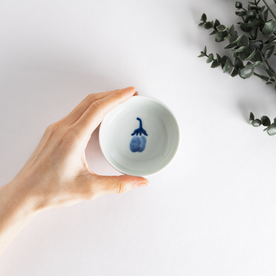 A small raised sauce dish featuring a minimalist blue eggplant design on the inside and vertical blue lines along the outer edge.