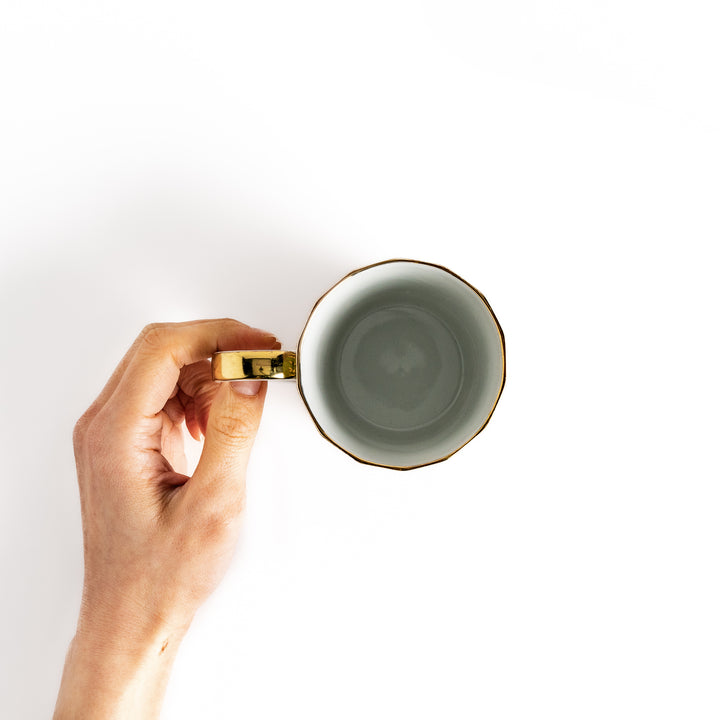 Hand holding a white mug with a gold rim and handle, viewed from above, showcasing its elegant design.