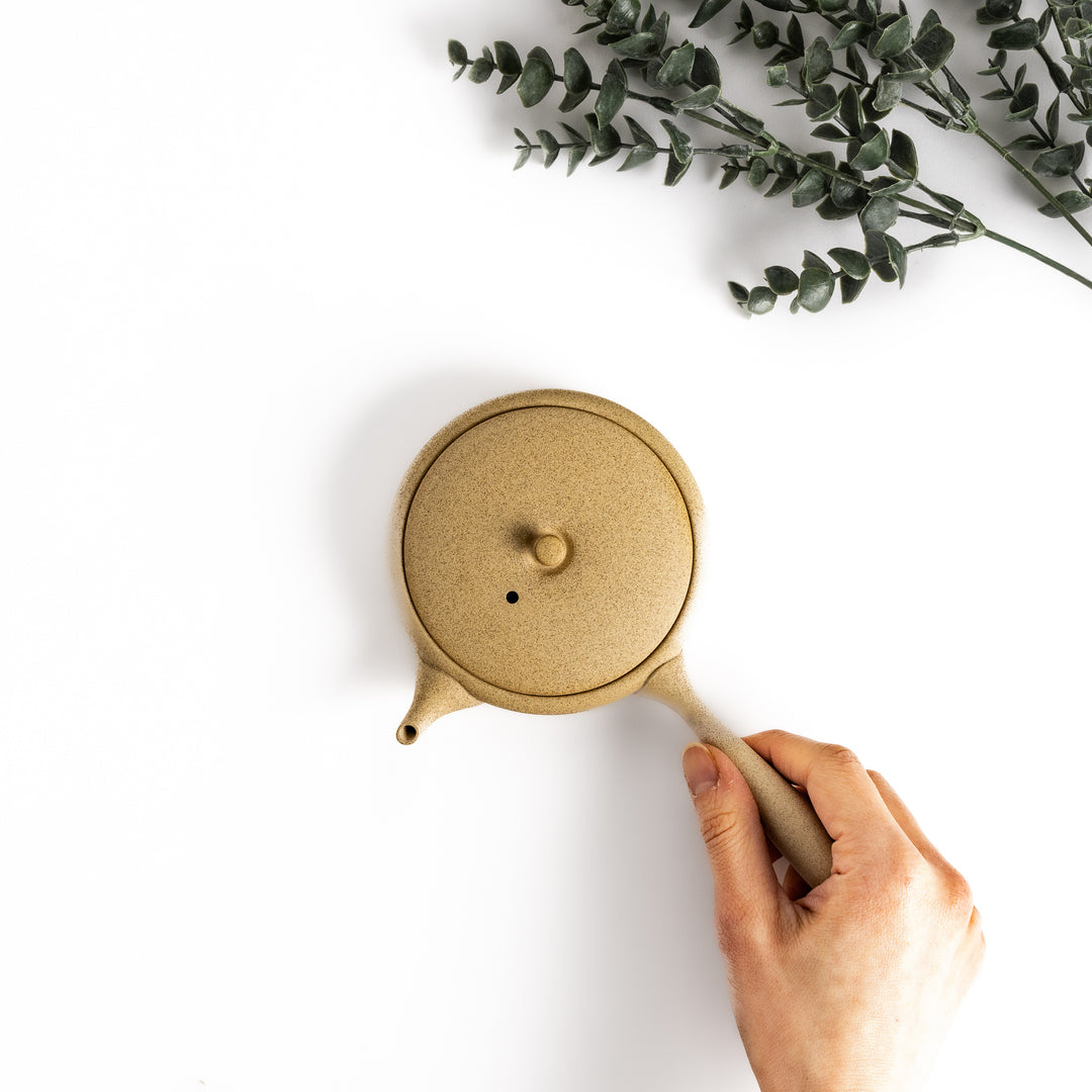 Hand-held beige teapot showcasing its flat shape and ergonomic handle, ideal for a refined tea-drinking experience.