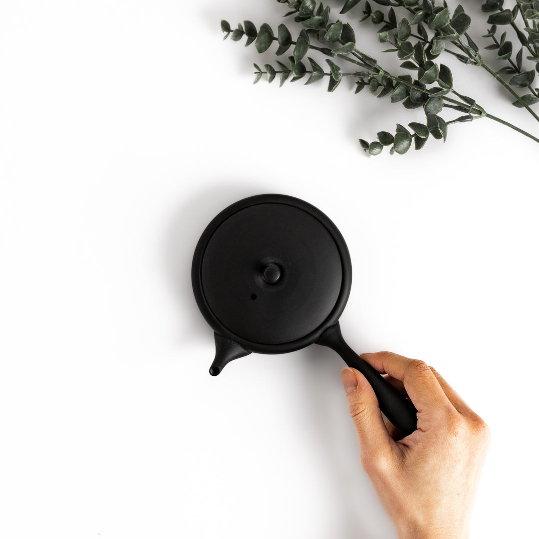 Hand-held black teapot showcasing its flat shape and ergonomic handle, ideal for a refined tea-drinking experience.