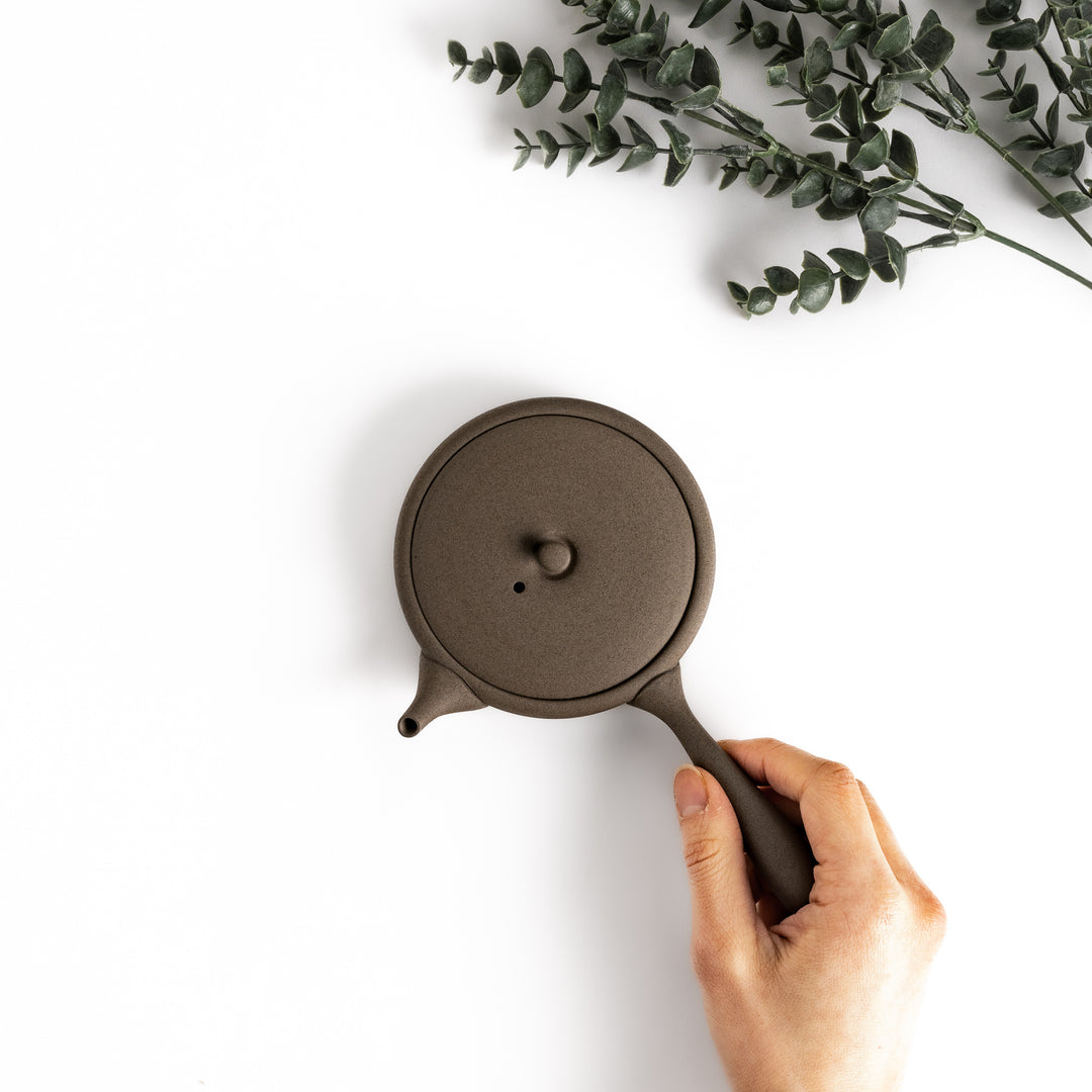 Hand-held gray teapot showcasing its flat shape and ergonomic handle, ideal for a refined tea-drinking experience.