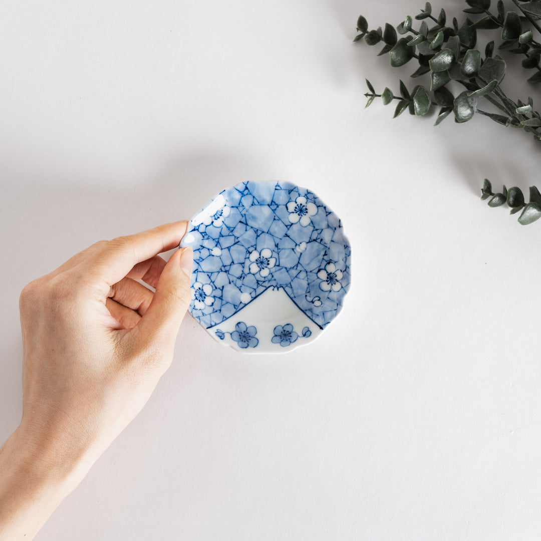 Hand holding a delicate round dish adorned with blue flowers and mountain designs.