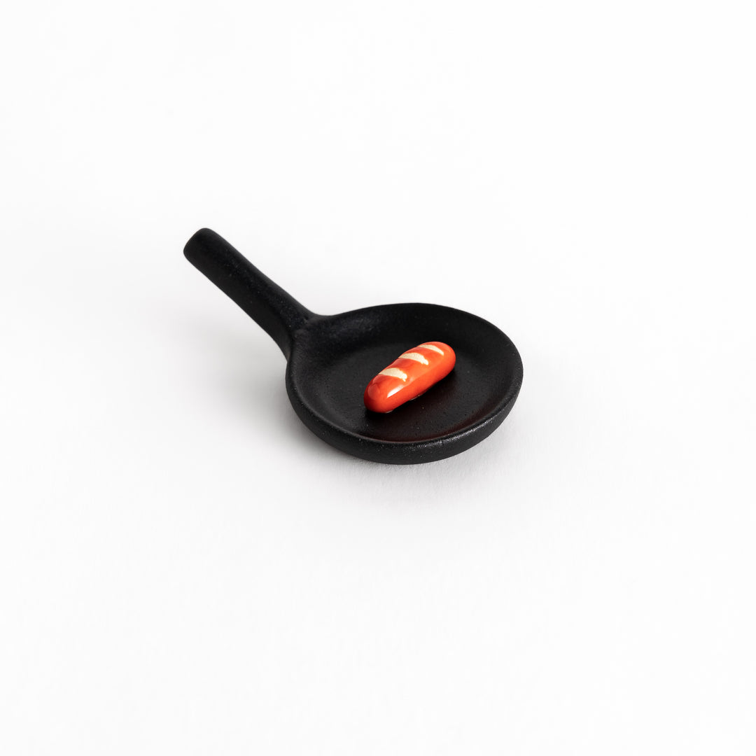 Black frying pan-shaped chopstick rest with a miniature red sausage detail, combining playful design with functionality.