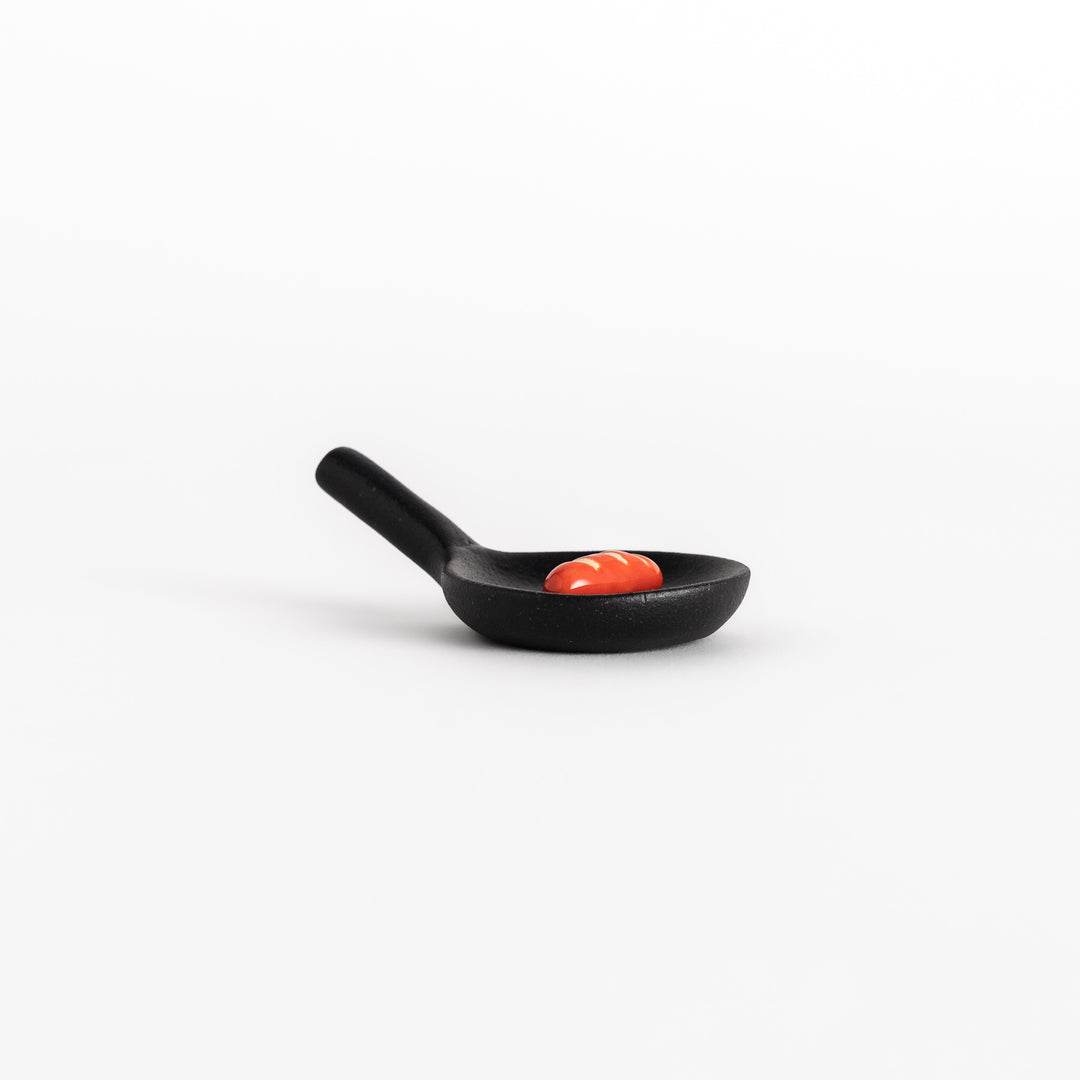 Side view of the black frying pan-shaped chopstick rest, showing its miniature sausage detail and functional handle.