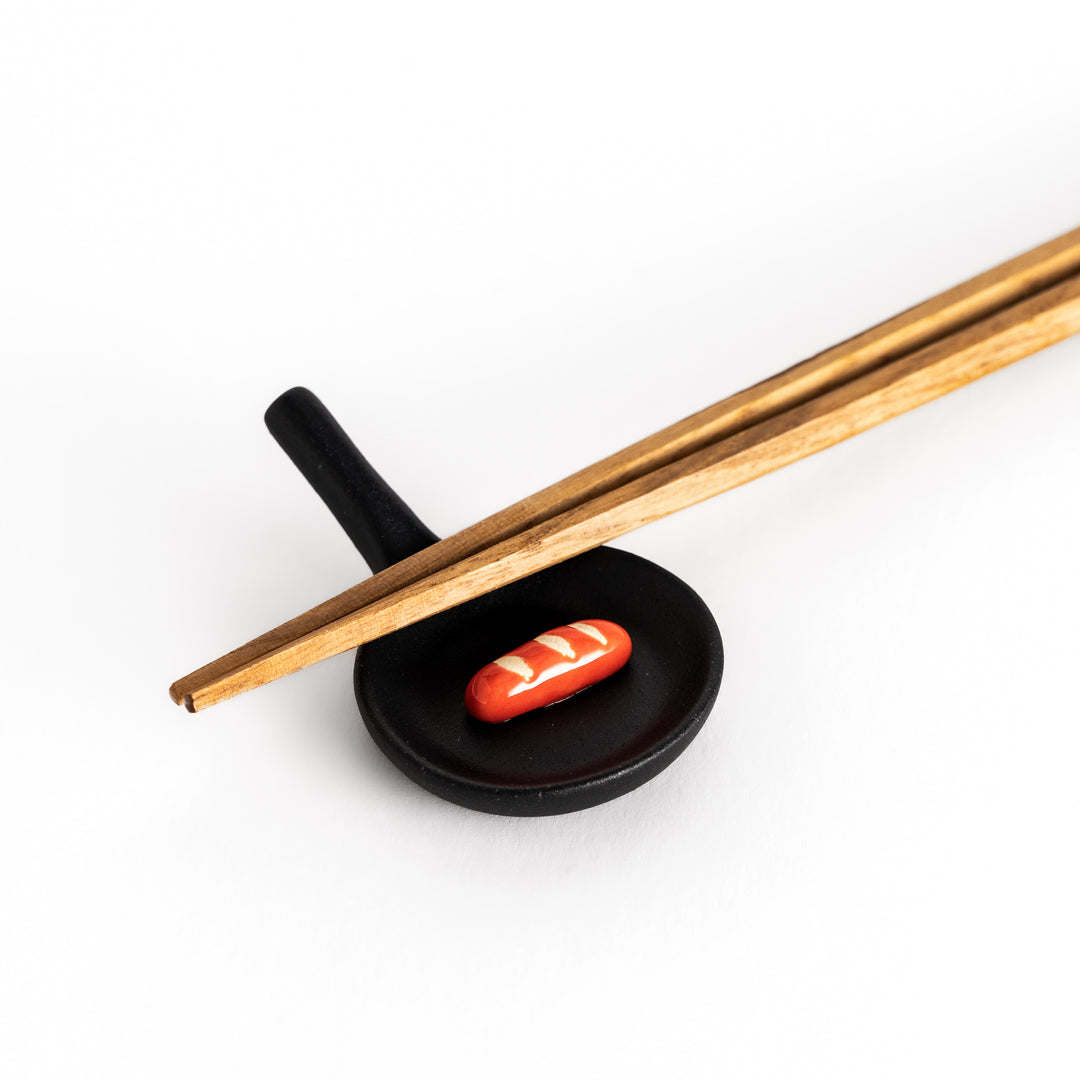 Close-up of a black frying pan-shaped chopstick rest with chopsticks, highlighting the miniature red sausage detail.