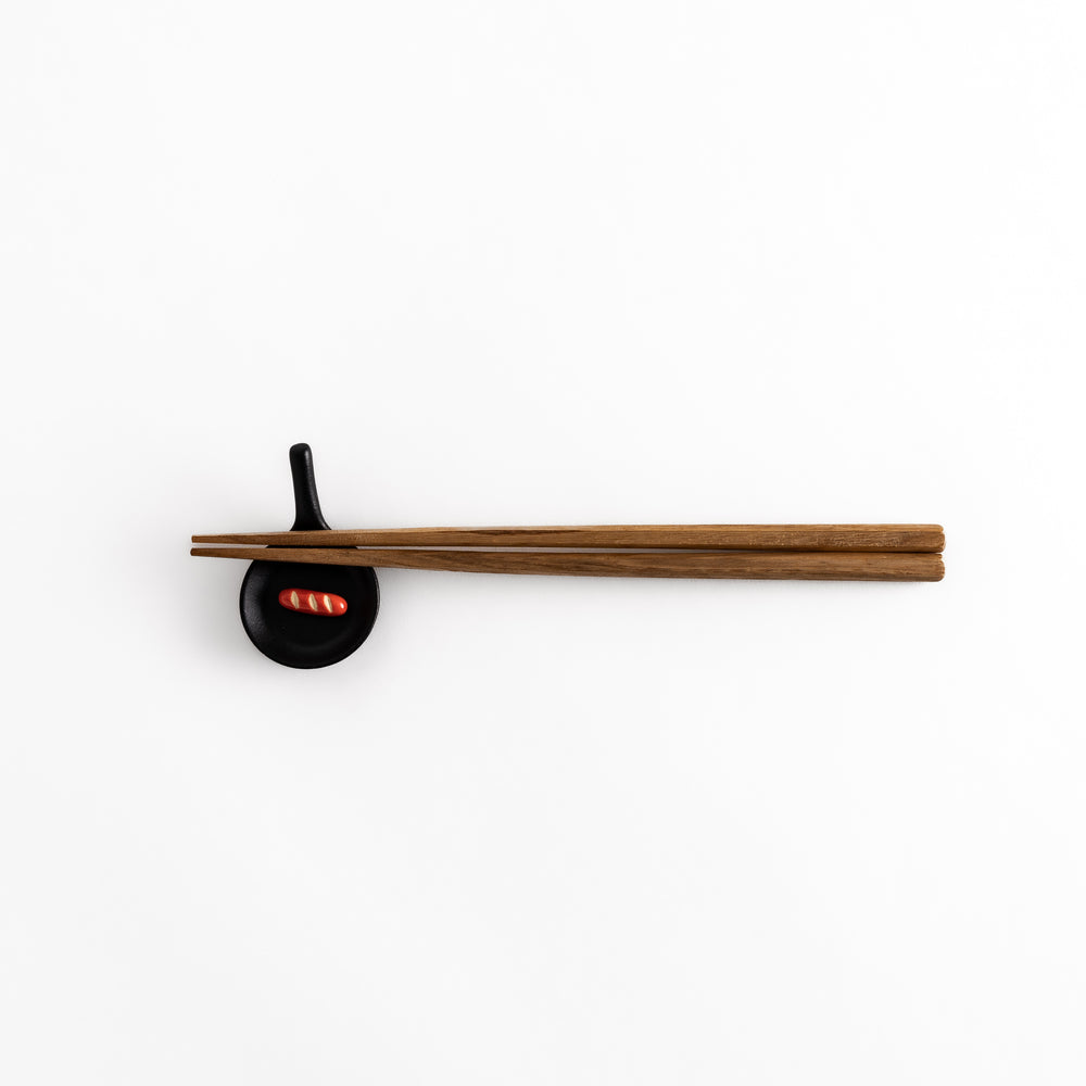 Pull-back view of a black frying pan-shaped chopstick rest with chopsticks resting on it, showcasing its practical use.