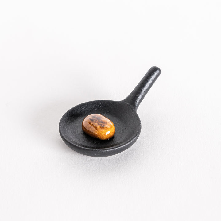 Slightly angled view of a small black frying pan chopstick rest, showing a realistic hamburger patty and chopsticks resting diagonally on top.
