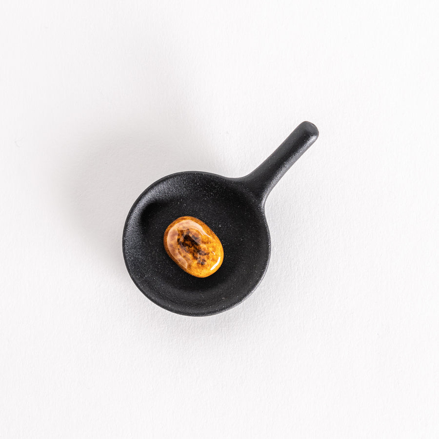 Top-down shot of a small black frying pan chopstick rest with a realistic-looking hamburger patty placed inside, captured on a clean white background.